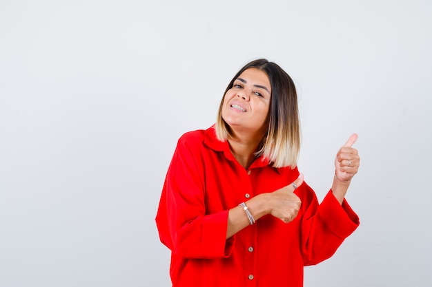 Foto linda mulher apontando para o lado com os polegares na blusa vermelha e parecendo alegre. vista frontal.