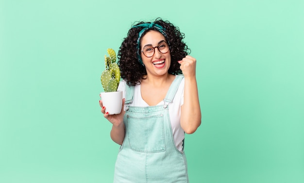 Linda mulher árabe chocada, rindo, comemorando o sucesso e segurando um cacto em um vaso