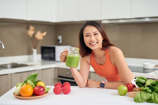 Linda mulher asiática bebendo suco desintoxicante verde enquanto usa roupas esportivas