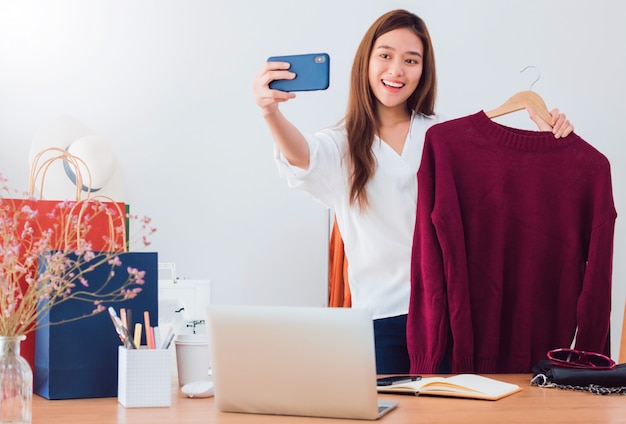 Linda mulher asiática blogueira está mostrando e analisa o produto. na frente da câmera do smartphone em casa.