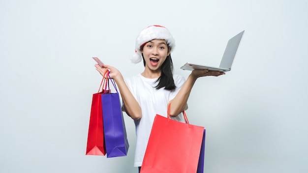 Linda mulher asiática com chapéu de Papai Noel segurando a sacola de compras e o laptop em estúdio um tiro.