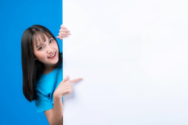 Linda mulher asiática com estilo de cabelo franja na camiseta azul sorrindo e apontando o dedo um espaço em branco para banner de publicidade espaço de cópia vazio quadro branco um banner em branco isolado em fundo azul