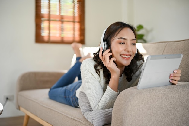 Linda mulher asiática com fones de ouvido assistindo filme através de seu tablet enquanto estava deitada no sofá