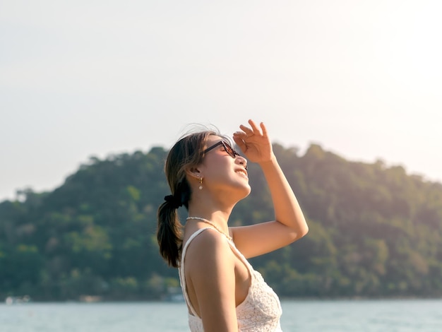 Linda mulher asiática com óculos de sol na praia em vibrações de verão Sorriso feliz fêmea cobrindo a luz do sol com a mão na ilha de montanha verde mar e fundo do céu Férias de férias