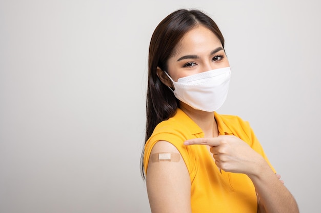 Linda mulher asiática de camisa amarela usando máscara recebendo uma proteção vacinal contra o coronavírus. Feliz feminino apontando o braço com curativo após receber a vacinação.