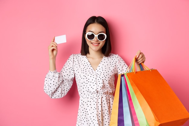 Linda mulher asiática de óculos escuros indo às compras, segurando sacolas e mostrando o cartão de crédito, em pé sobre a rosa