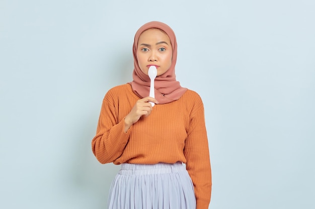 Linda mulher asiática de suéter marrom e hijab cobrindo a boca com colher quer experimentar comida deliciosa isolada em fundo branco