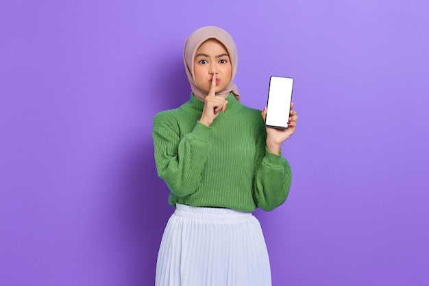 Linda mulher asiática de suéter verde e hijab mostrando celular de tela em branco e fazendo gesto de silêncio isolado sobre fundo roxo