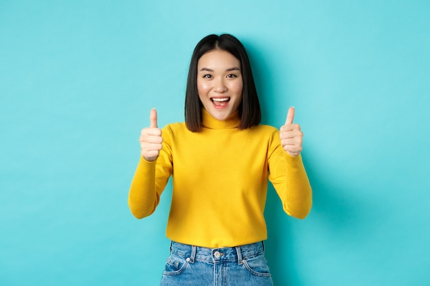 Linda mulher asiática elogia o bom trabalho, mostrando os polegares para cima gesto e sorrindo em aprovação, recomenda o produto, satisfeita em pé sobre o fundo azul.