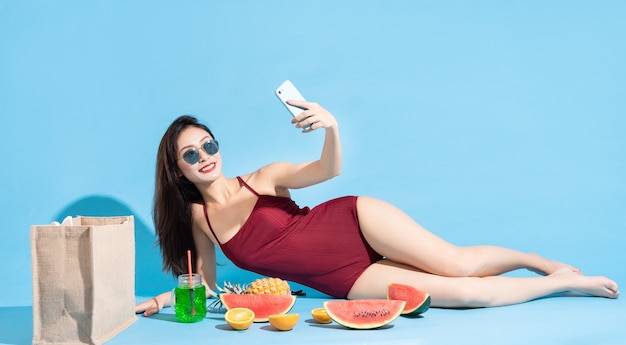 Linda mulher asiática em biquíni vermelho, mentindo e usando o telefone para tirar uma selfie com frutas tropicais ao redor. conceito de viagens de praia tropical no verão