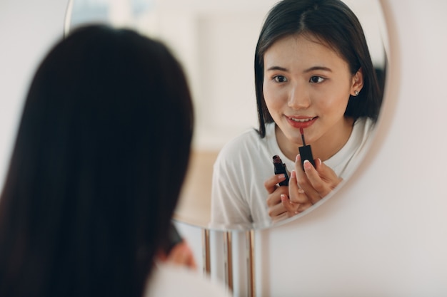 Linda mulher asiática em frente ao espelho aplicando maquiagem nos lábios com batom na sala de estar