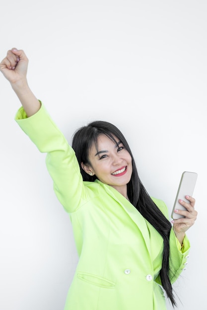 Linda mulher asiática em pastel de terno verde usando telefone inteligente gesticulando sim isolado no fundo branco Garota alegre feliz com cabelos longos comemorando a vitória nas mídias sociais de sucesso da mulher asiática
