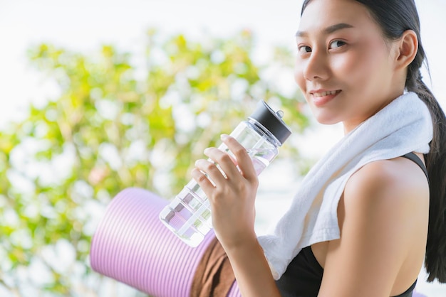 Linda mulher asiática esportiva segurando tapete de ioga e garrafa de água para treino de ioga de fitness saudável com fundo de jardim