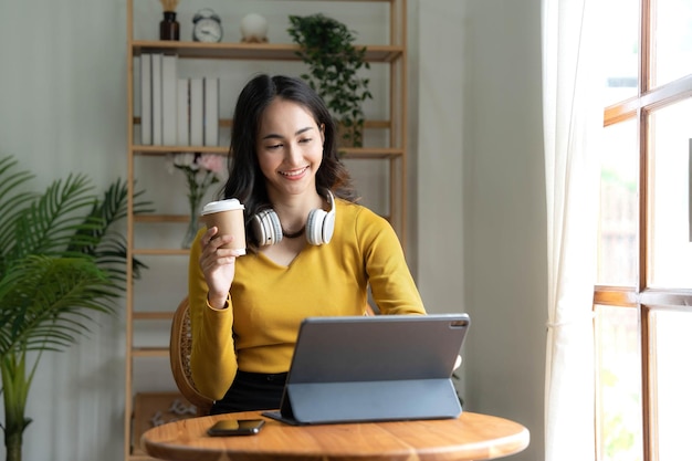 Linda mulher asiática está usando laptop bebendo café e sorrindo enquanto está sentado perto da janela em casa