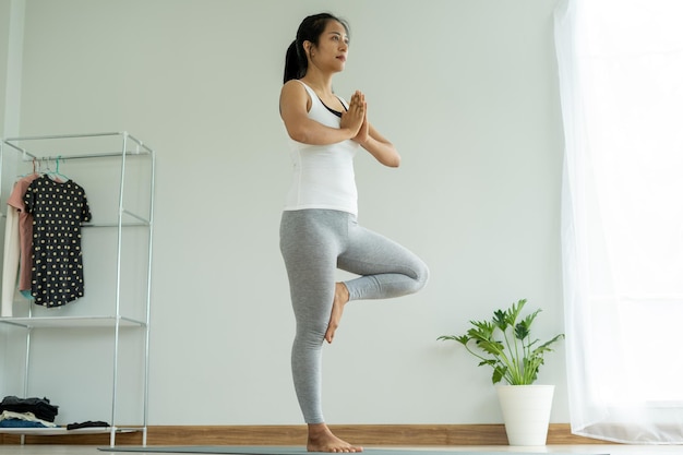 Linda mulher asiática fazendo ioga em casa as mulheres mantêm o equilíbrio em uma pose de árvore e praticam a inspiração e a expiração lentamente conceito de saúde e fitness