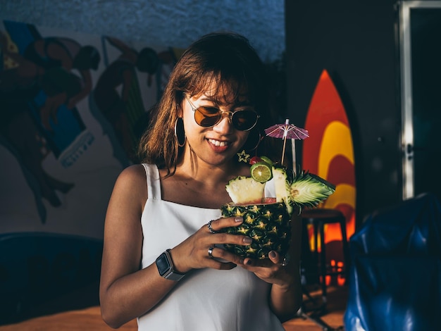 Linda mulher asiática feliz vestindo branco sem mangas e óculos de sol segurando coquetel de suco de abacaxi no verão