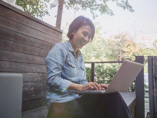 Linda mulher asiática feliz vestindo camisa jeans azul usando computador portátil no colo em fundo de madeira