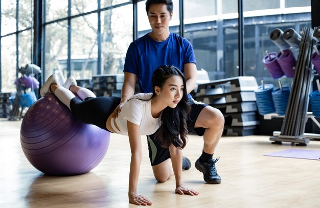 Linda mulher asiática jogando ioga por bola de ioga com o homem treinador no ginásio no conceito de exercício no ginásio. jovens casais com exercícios fazendo ioga juntos no interior.