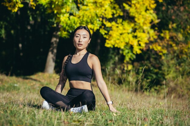 Linda mulher asiática medita na floresta em um dia ensolarado, o atleta descansa sentado na posição de lótus e olha feliz e calmamente para a câmera