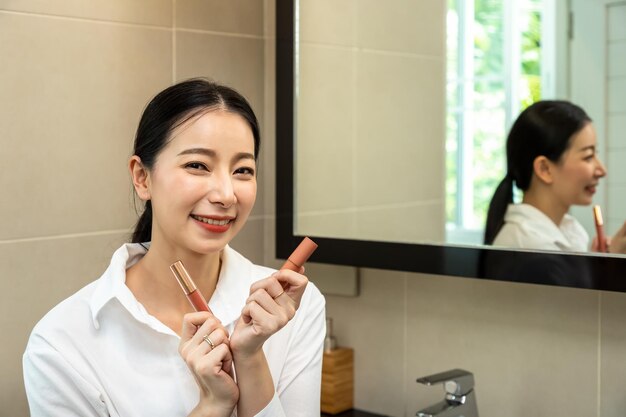 Linda mulher asiática na frente do espelho e comparar batom de maquiagem labial no banheiro do banheiro em pó