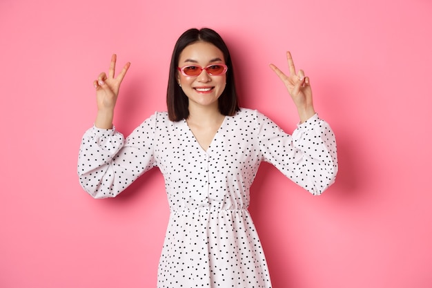 Linda mulher asiática nas férias de verão, mostrando sinais de paz e sorrindo feliz, usando óculos escuros da moda, rosa.