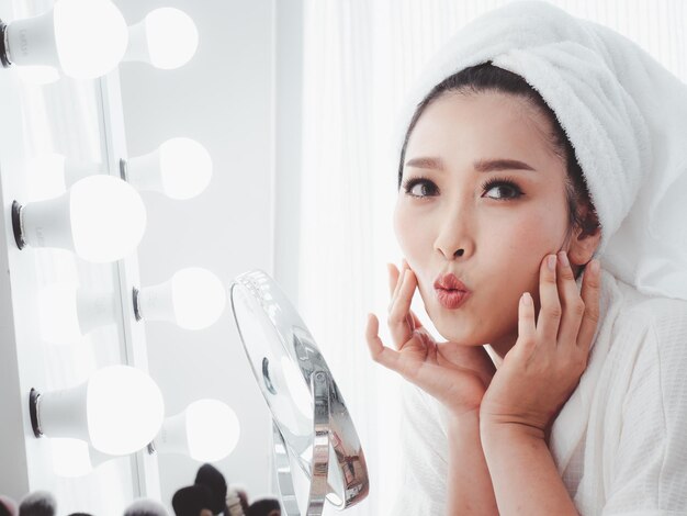 Linda mulher asiática olhando no espelho e aplicando cosméticos com um pincel pequeno Mulher atraente aplicando sua maquiagem refletida em um espelho com uma lâmpada surround em homexA
