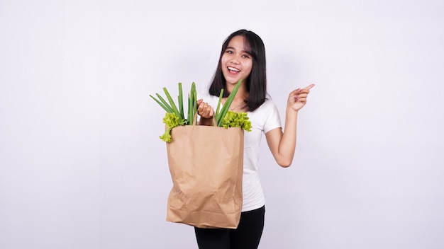 Linda mulher asiática segurando um saco de papel com legumes frescos e muito feliz apontando com a mão e o dedo na superfície branca isolada
