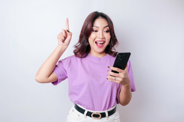 Linda mulher asiática sorridente apontando o dedo para cima para o espaço vazio enquanto segura seu telefone isolado de fundo branco