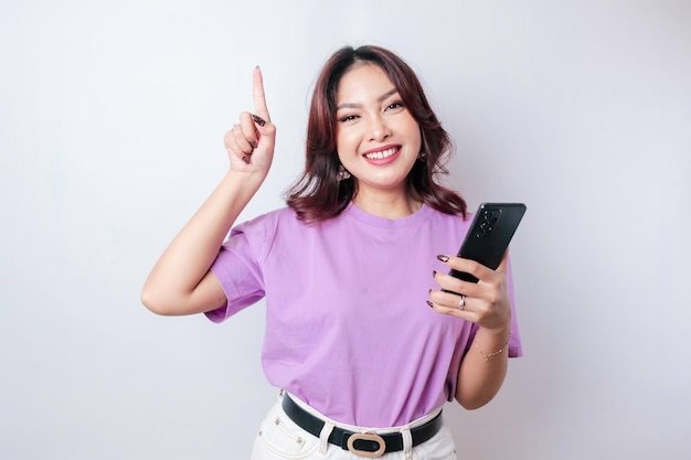 Linda mulher asiática sorridente apontando o dedo para cima para o espaço vazio enquanto segura seu telefone isolado de fundo branco