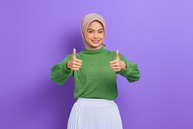 Linda mulher asiática sorridente com suéter verde mostrando gesto de polegar para cima com a mão isolada sobre o fundo roxo