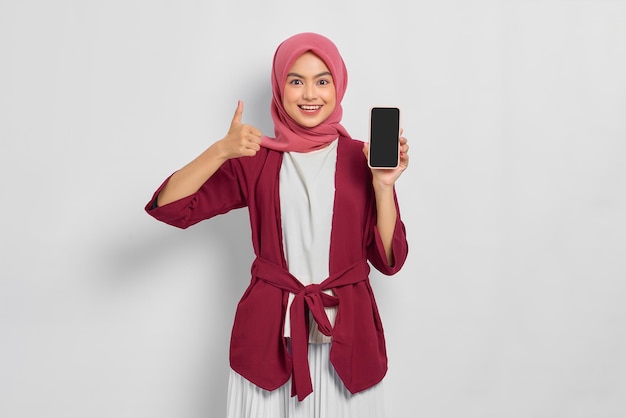 Linda mulher asiática sorridente em camisa casual mostrando a tela em branco do celular e gesticulando polegares para cima isolados sobre fundo branco