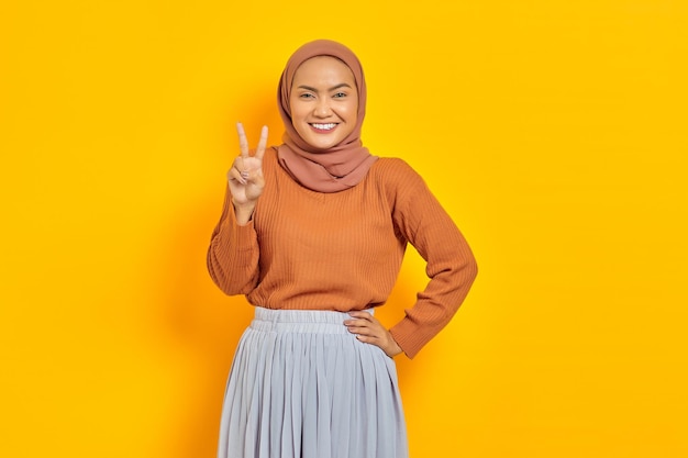 Linda mulher asiática sorridente em suéter marrom e hijab olhando para a câmera e mostrando sinal de paz com o dedo isolado sobre fundo amarelo. conceito de estilo de vida religioso de pessoas