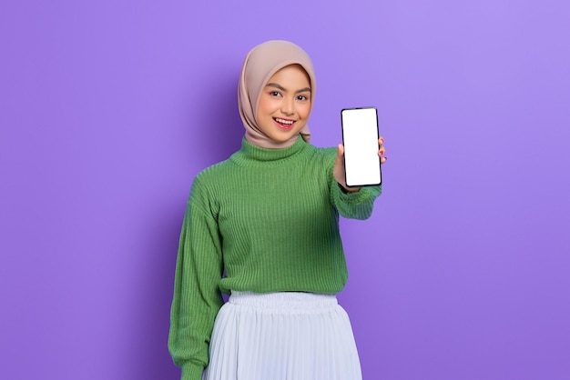 Linda mulher asiática sorridente em suéter verde e hijab mostrando celular de tela em branco isolado sobre fundo roxo