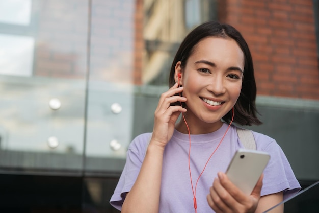 Linda mulher asiática sorridente segurando celular ouvindo música na rua olhando para a câmera