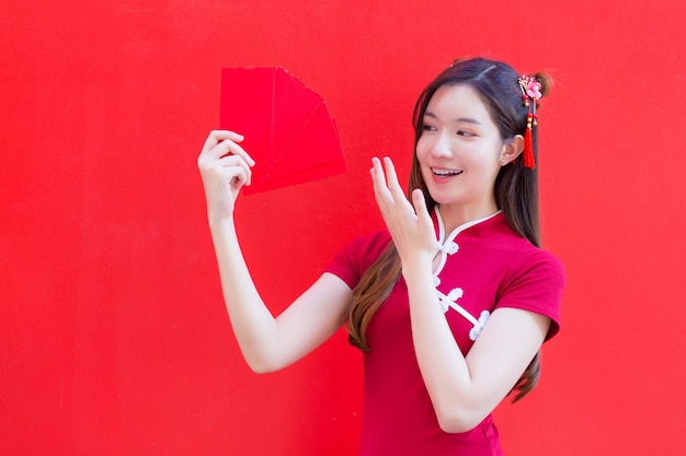 Linda mulher asiática usa um cheongsam vermelho e segura envelopes vermelhos enquanto olha para a câmera