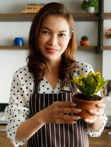 Linda mulher asiática usando avental segurando e mostrando pequenas plantas e flores no jardim doméstico com um sorriso e uma cara feliz. Hobby e conceito de trabalho de tempo livre.