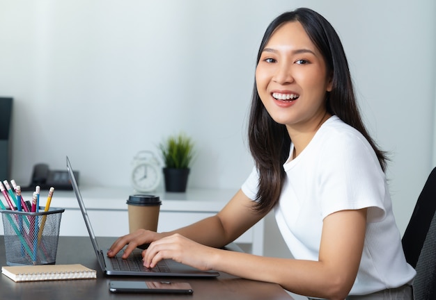 Linda mulher asiática usando laptop e sentado à mesa no escritório em casa.