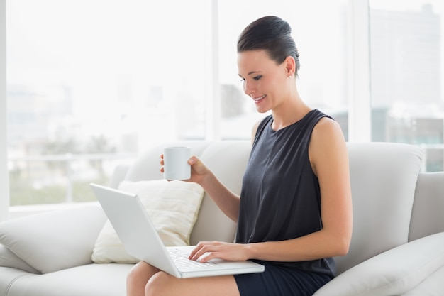 Linda mulher bem vestida usando laptop enquanto toma café no sofá