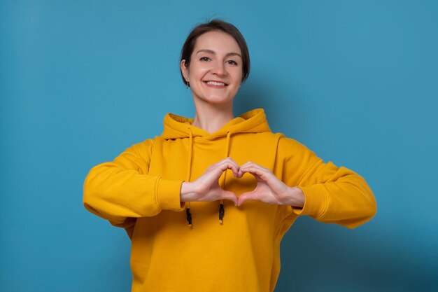 Linda mulher caucasiana com sorriso feliz molda o coração com as duas mãos expressa amor por você Estúdio filmado na parede azul