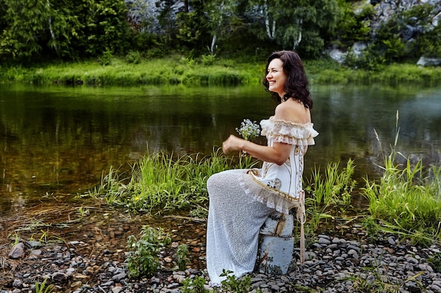 Linda mulher caucasiana em uma noite ou vestido de noiva senta-se nas margens do rio selvagem com buquê de flores silvestres nas mãos, ela ri.