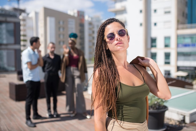 Linda mulher caucasiana na festa do telhado do terraço. Mulher de cabelos compridos em roupas casuais e óculos escuros. Pessoas de diferentes nacionalidades em pé no fundo. Teambuilding, conceito de festa