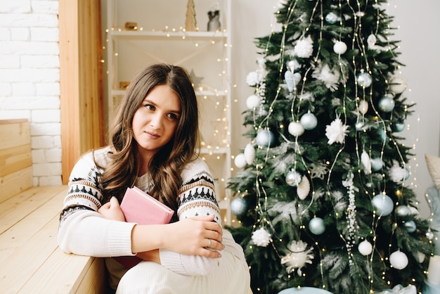 Linda mulher caucasiana sentada ao lado de uma linda árvore de Natal decorada segurando um livro e sonhando