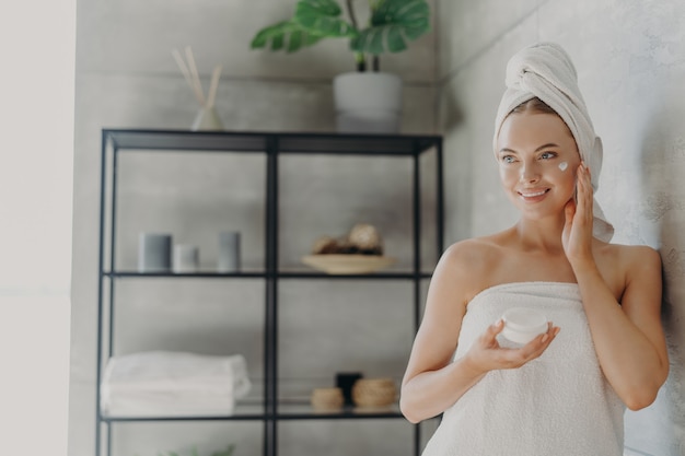 Linda mulher caucasiana sorridente aplica creme de beleza na bochecha, desfruta de rotina matinal de cuidados com a pele doméstica, enrolada em toalha de banho, arrumando-se após tomar banho em poses no banheiro conceito de higiene