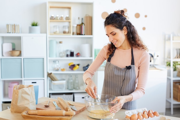 Linda mulher caucasiana usando avental em pé à mesa na cozinha moderna batendo ovos em uma tigela de vidro com o chicote