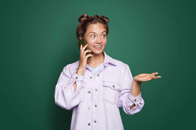 Foto linda mulher caucasiana, vestindo uma camisa moderna, falando ao telefone e posando em uma parede verde