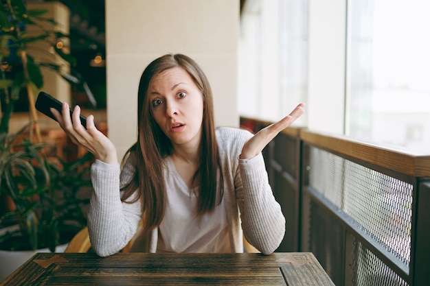 Linda mulher chateada sentada sozinha perto da grande janela na cafeteria, relaxando no restaurante durante o tempo livre. Mulher triste tendo uma conversa com o celular, descanse no café. Conceito de estilo de vida.