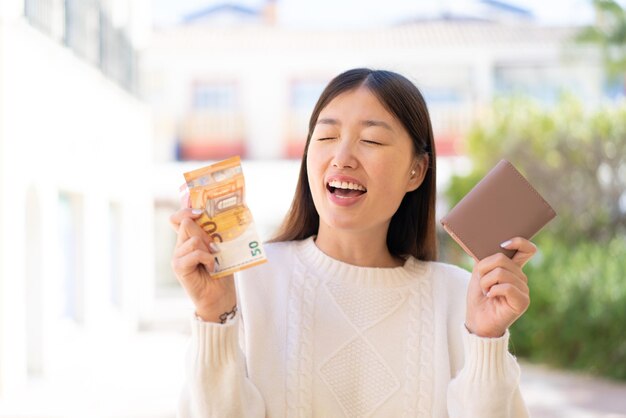 Foto linda mulher chinesa ao ar livre segurando carteira com dinheiro com expressão feliz