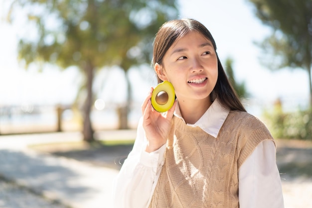 Linda mulher chinesa ao ar livre segurando um abacate com expressão feliz