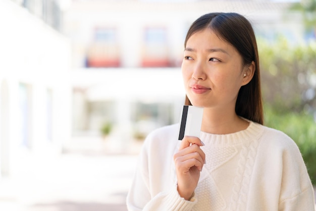 Linda mulher chinesa ao ar livre segurando um cartão de crédito e pensando