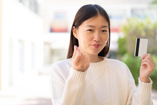 Linda mulher chinesa ao ar livre segurando um cartão de crédito fazendo gesto de dinheiro